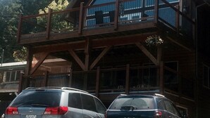 Uma cabana de madeira de dois andares com varanda, carros estacionados em frente e chaminé de pedra.