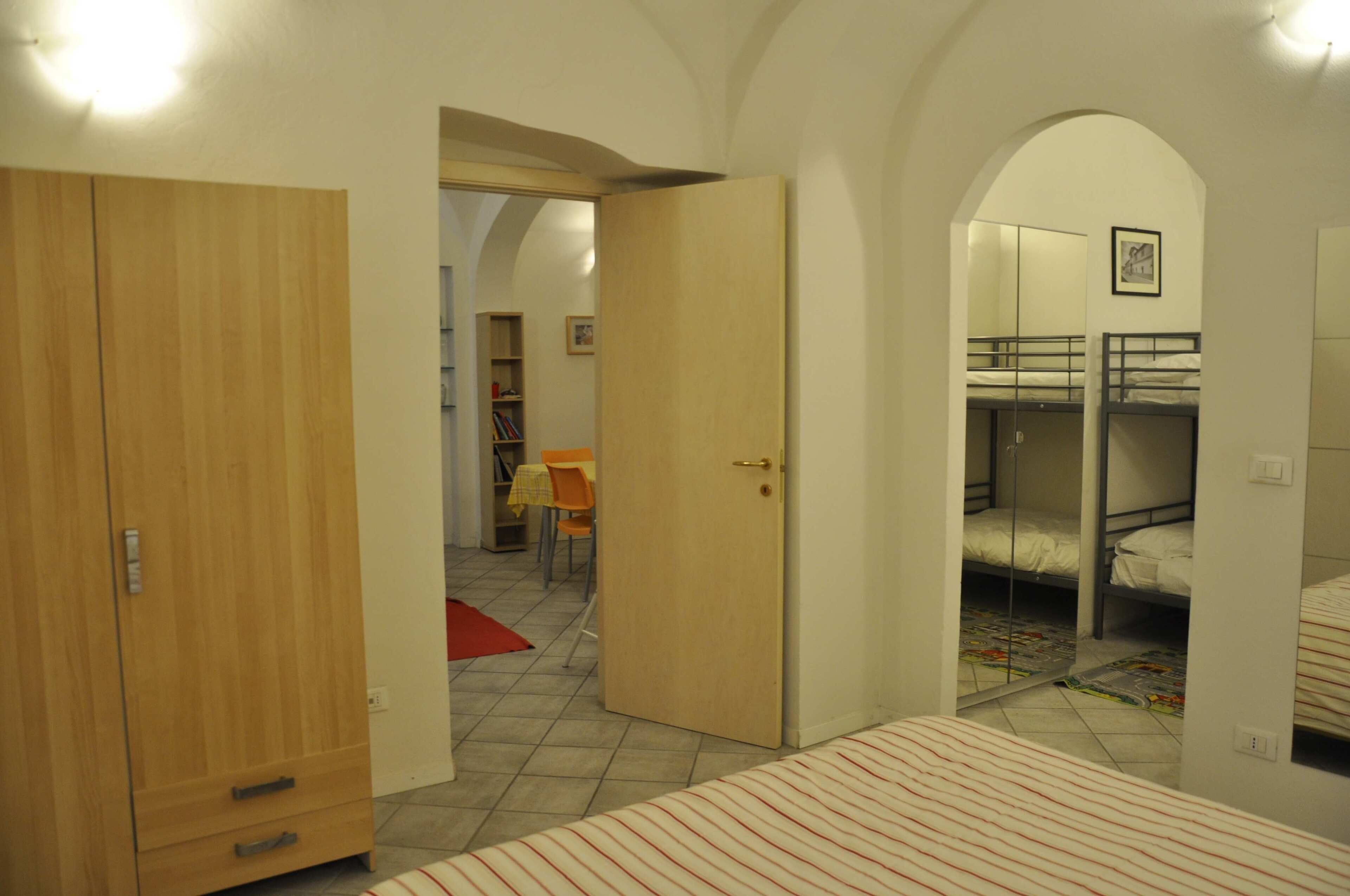 Una stanza con letti a castello, un armadio in legno e una porta che dà su un'altra stanza con una sedia e un tavolo.