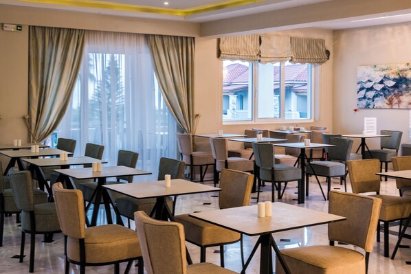 Un comedor con varias mesas y sillas, ventanas grandes con cortinas, y un cuadro en la pared.