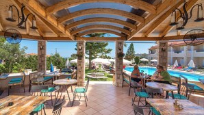 An outdoor patio area with wooden beams and tables, overlooking a swimming pool. - Windmill Hotel (Zakynthos)