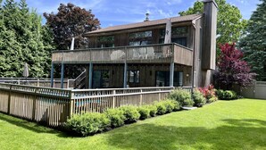 Une maison en bois à deux étages, dotée d’un balcon, entourée d’une pelouse bien entretenue et de divers arbustes.