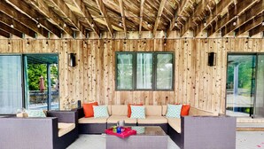Une terrasse en bois avec un canapé et deux petites tables d’appoint, de grandes fenêtres et une vue sur la verdure.