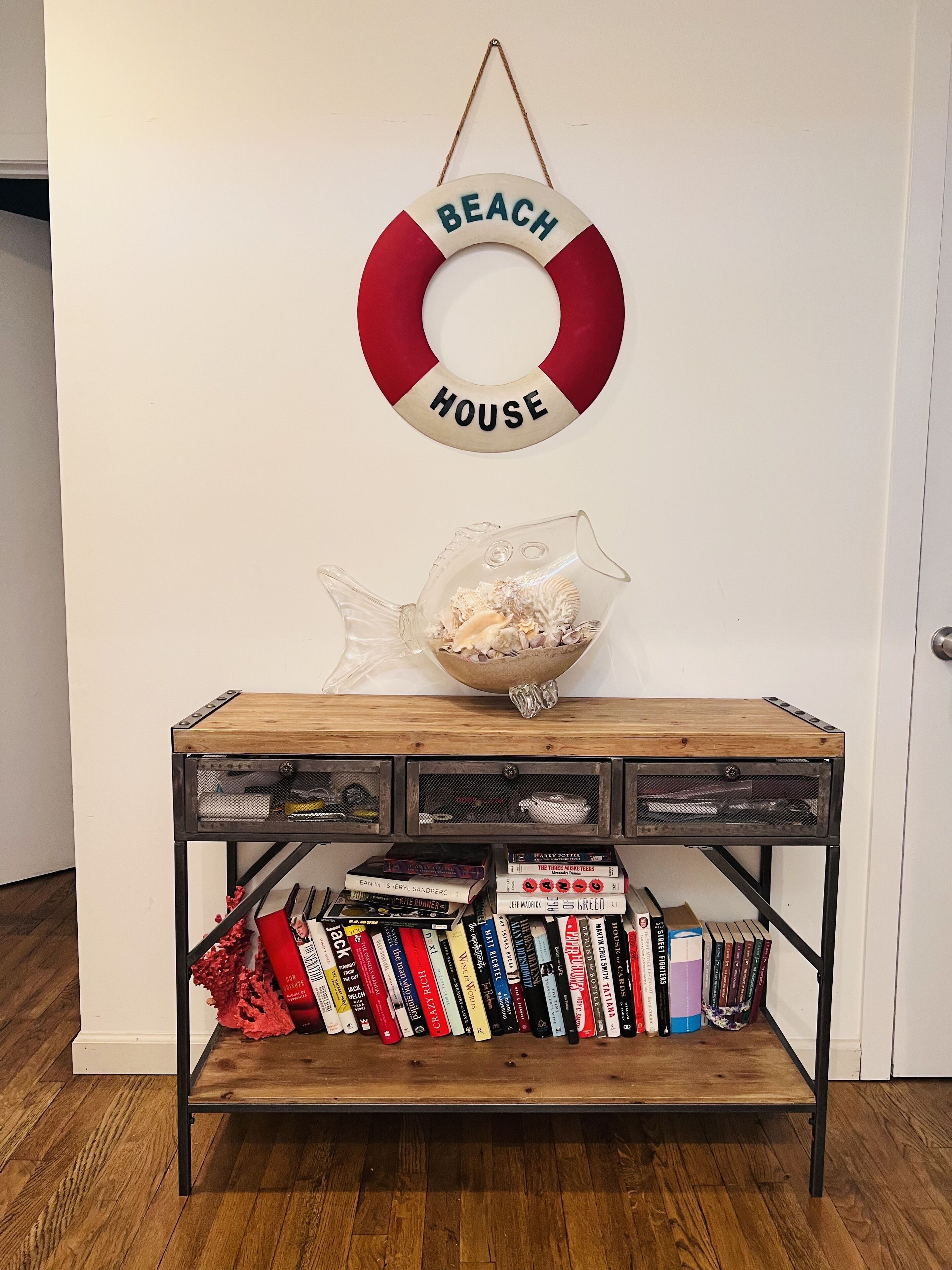 Una mesa de madera con un adorno de chaleco salvavidas, un cuenco de vidrio con conchas marinas y una estantería con varios libros.
