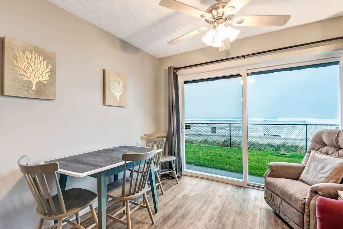 A dining area with a table and chairs, a ceiling fan, and a large sliding glass door leading to a grassy outdoor area with a view of the ocean.