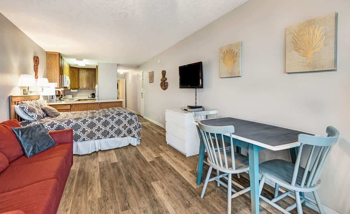 A compact room with a bed, a red sofa, a dining table with two chairs, a small kitchenette, and a TV.