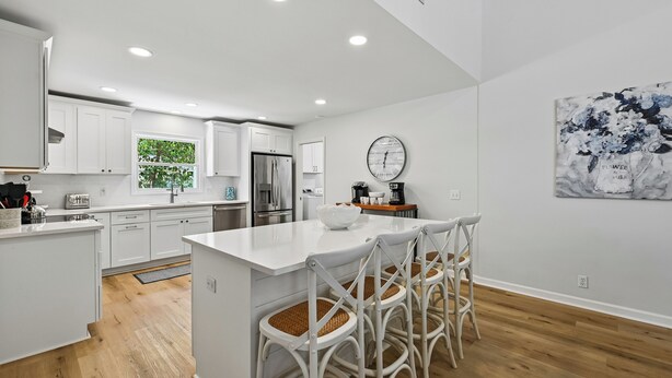 Una cocina moderna con armarios blancos, una isla central con taburetes y un refrigerador de acero inoxidable, además de un reloj en la pared.