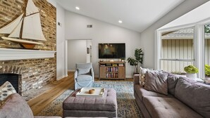 Una sala de estar con chimenea de ladrillo, un televisor grande, un sillón gris, una estantería y una ventana con plantas.