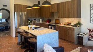 A modern kitchen with wooden cabinets, a stainless steel refrigerator, a central island with bar stools, pendant lights, and a dining area with a round table and chairs. - Relax in the Best and Biggest Space in Palm Valley Country Club. (Palm Desert)