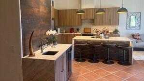 A modern kitchen with a central island, bar stools, pendant lighting, and a mix of wood and stone finishes. - Relax in the Best and Biggest Space in Palm Valley Country Club. (Palm Desert)