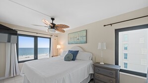 Ein Schlafzimmer mit Bett, Nachttisch, Deckenventilator, Fenster mit Blick auf den Strand und einem Bild an der Wand.