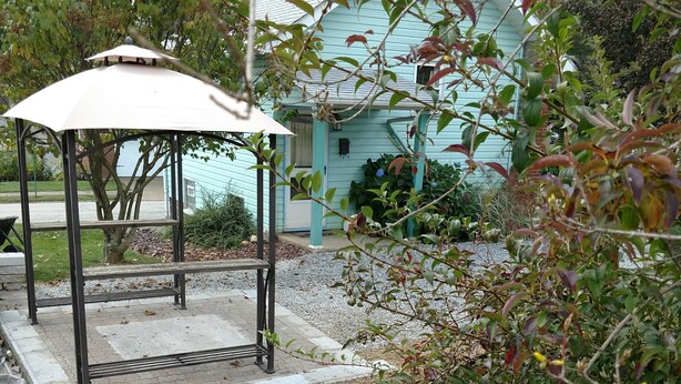 Un porche con un banco, rodeado de un jardín con diversas plantas y una casa al fondo.