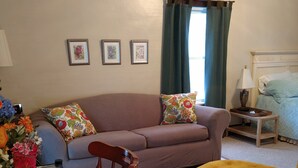 Una sala de estar con un sofá, un reposapiés amarillo, una silla de madera, una mesita y una ventana con cortinas.
