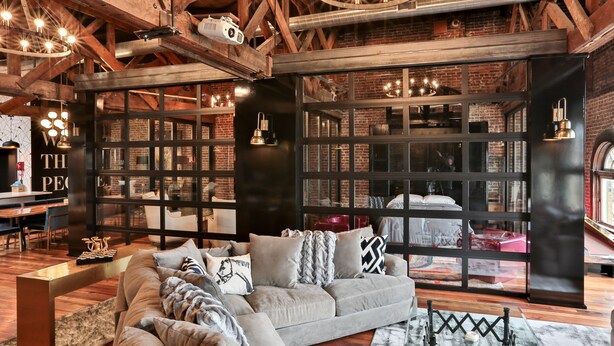 A cozy living room with a large sectional sofa, a coffee table, and a fireplace. There are bookshelves filled with books and decorative items. The room has exposed wooden beams and a brick wall.