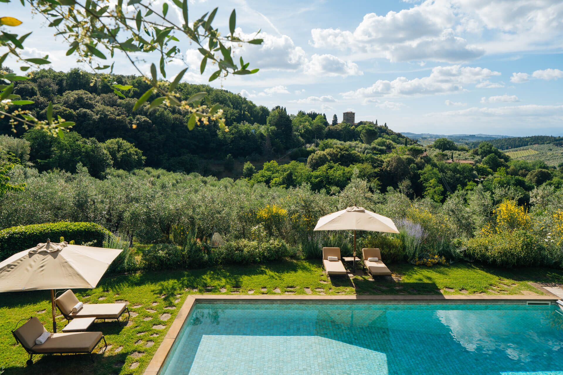 A pool with lounge chairs and umbrellas, surrounded by lush greenery and hills.