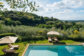 Una piscina con sillas de descanso y sombrillas, rodeada de abundante vegetación y colinas.