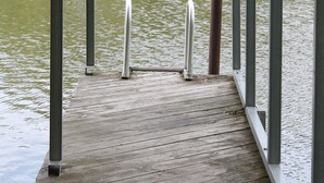 Ein hölzerner Steg erstreckt sich über einen stillen See, im Hintergrund sieht man ein Haus.