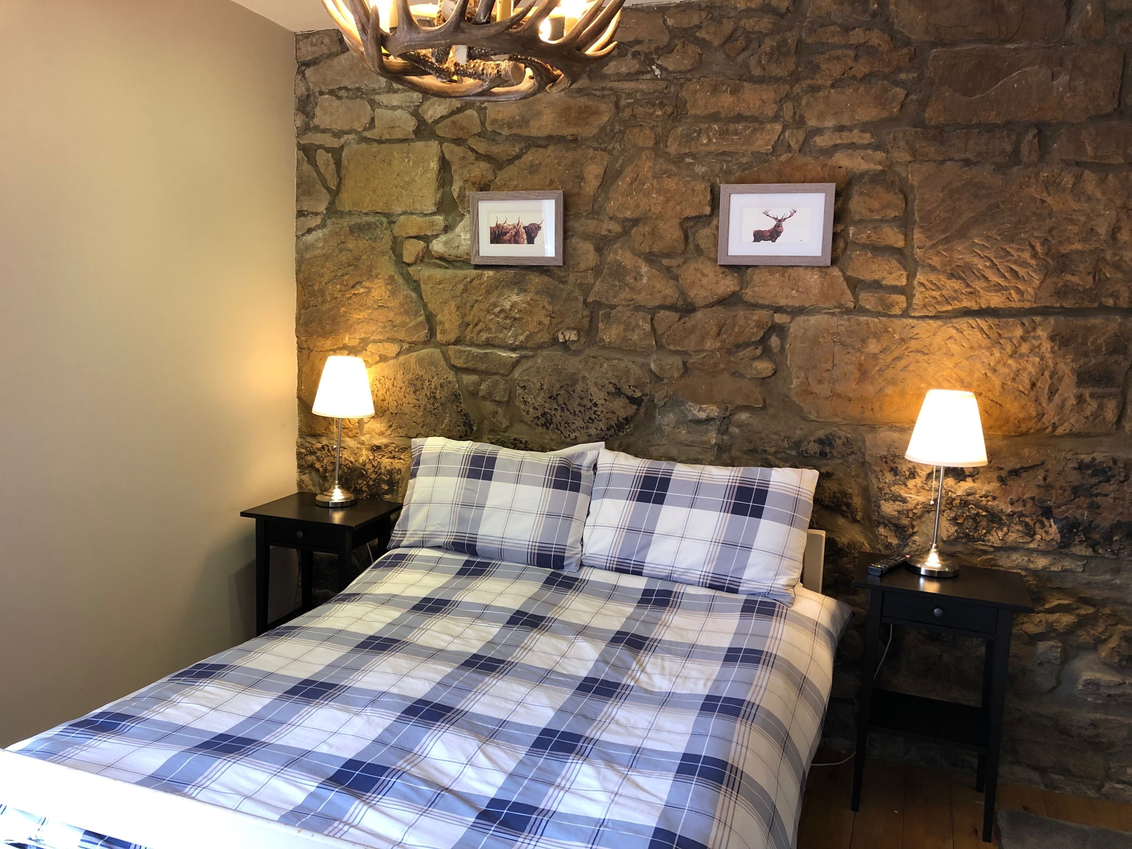A bedroom with a stone wall, a bed with a plaid bedspread, two bedside tables with lamps, and framed pictures on the wall.