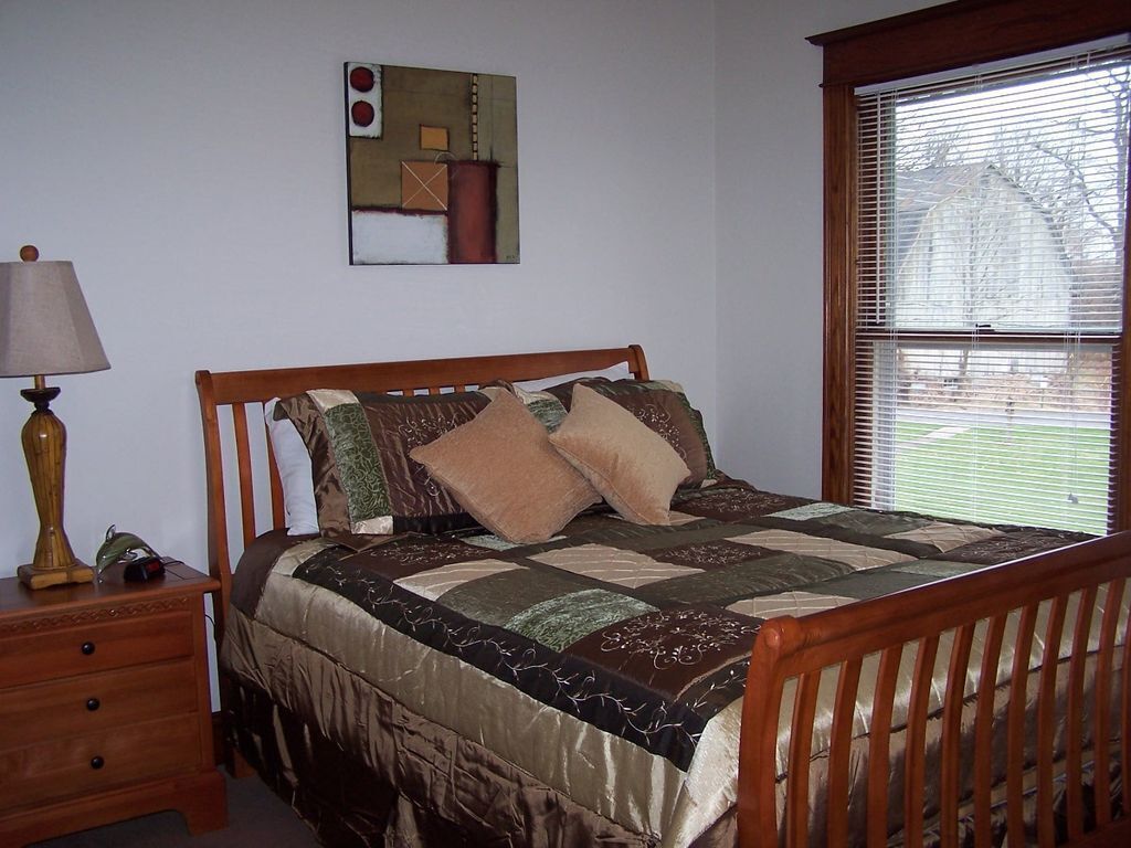 Une chambre à coucher avec un lit en bois, une table de chevet avec une lampe, un tableau au mur et une fenêtre avec des stores.