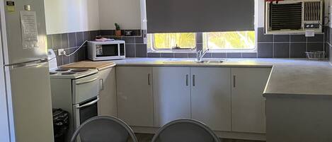 Eine kleine Küche mit Kühlschrank, Mikrowelle, Herd und Spüle. Zwei Stühle stehen an einem Tisch, und ein Fenster mit Jalousien ist vorhanden.