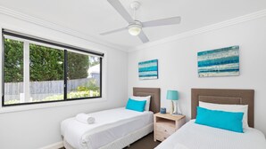 Habitación de hotel con dos camas, ventilador de techo, ventana con vista a vegetación y dos cuadros colgados en la pared.