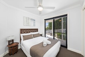 Un dormitorio con cama, ventilador de techo, mesita de noche, lámpara y vistas a través de un ventanal corredero.