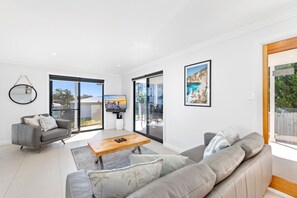 A modern living room with a grey sofa, a coffee table, and a TV mounted on the wall. There are large windows and a sliding glass door leading to an outdoor area.