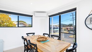 Uma área de refeições com mesa e cadeiras de madeira, um cesto trançado e vista para uma área residencial por grandes janelas.
