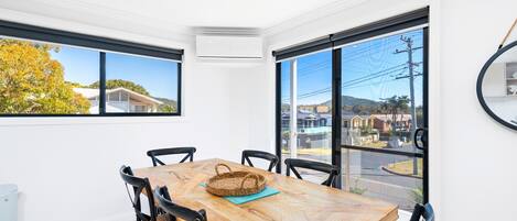 Uma área de refeições com mesa e cadeiras de madeira, um cesto trançado e vista para uma área residencial por grandes janelas.