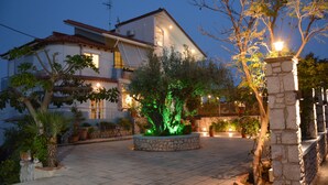 Une maison avec une terrasse carrelée, une fontaine en pierre et un arbre décoré de guirlandes lumineuses.