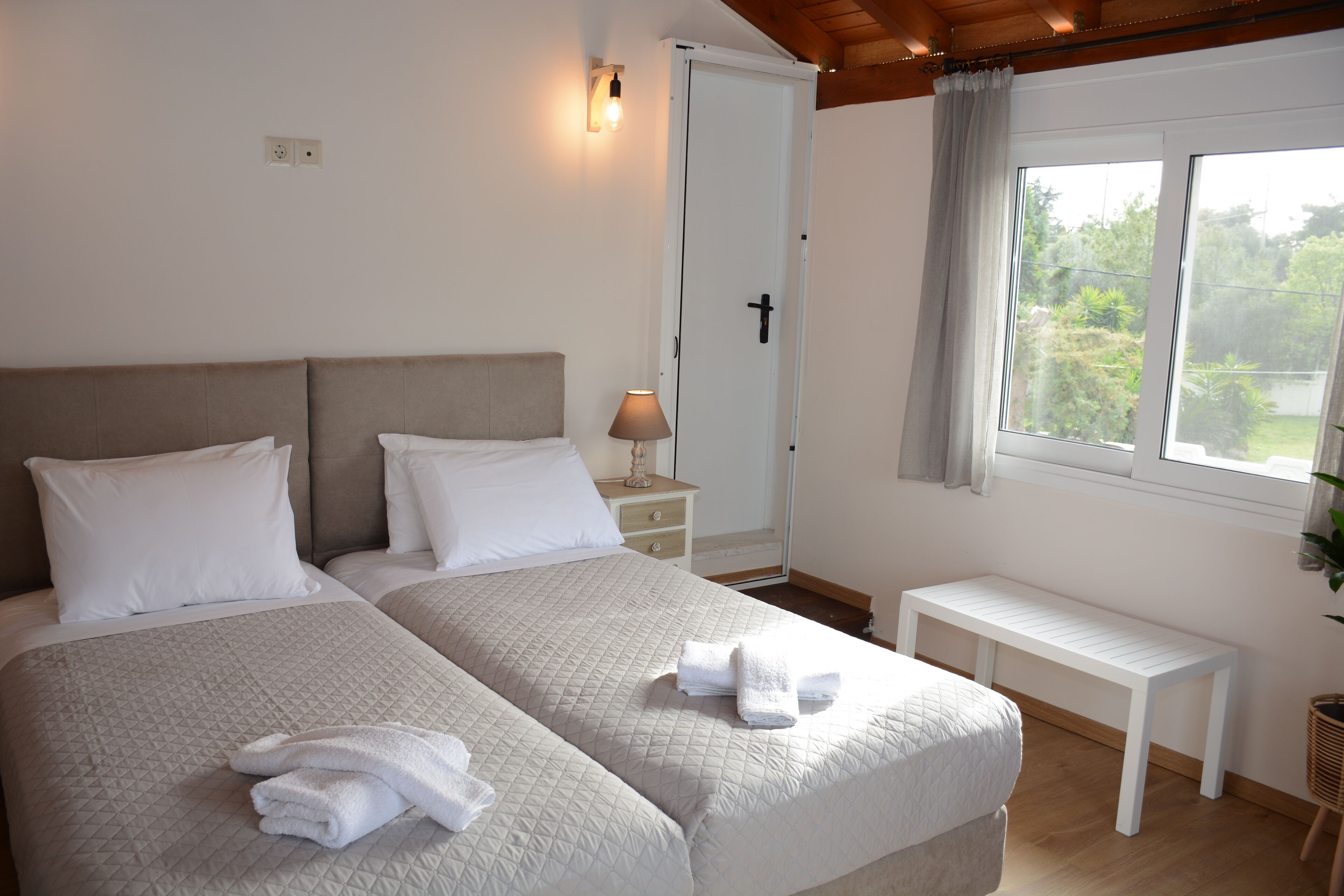 Un dormitorio con dos camas, una mesita de noche con lámpara, un banco y una ventana con vistas a zonas verdes.