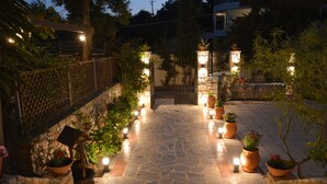 Un chemin de pierre menant à une entrée fermée par un portail, bordé de plantes en pot et éclairé par des lampes fixées aux murs.