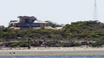 A coastal house with a large balcony overlooking the sea, a wind turbine in the background, and a rocky landscape.