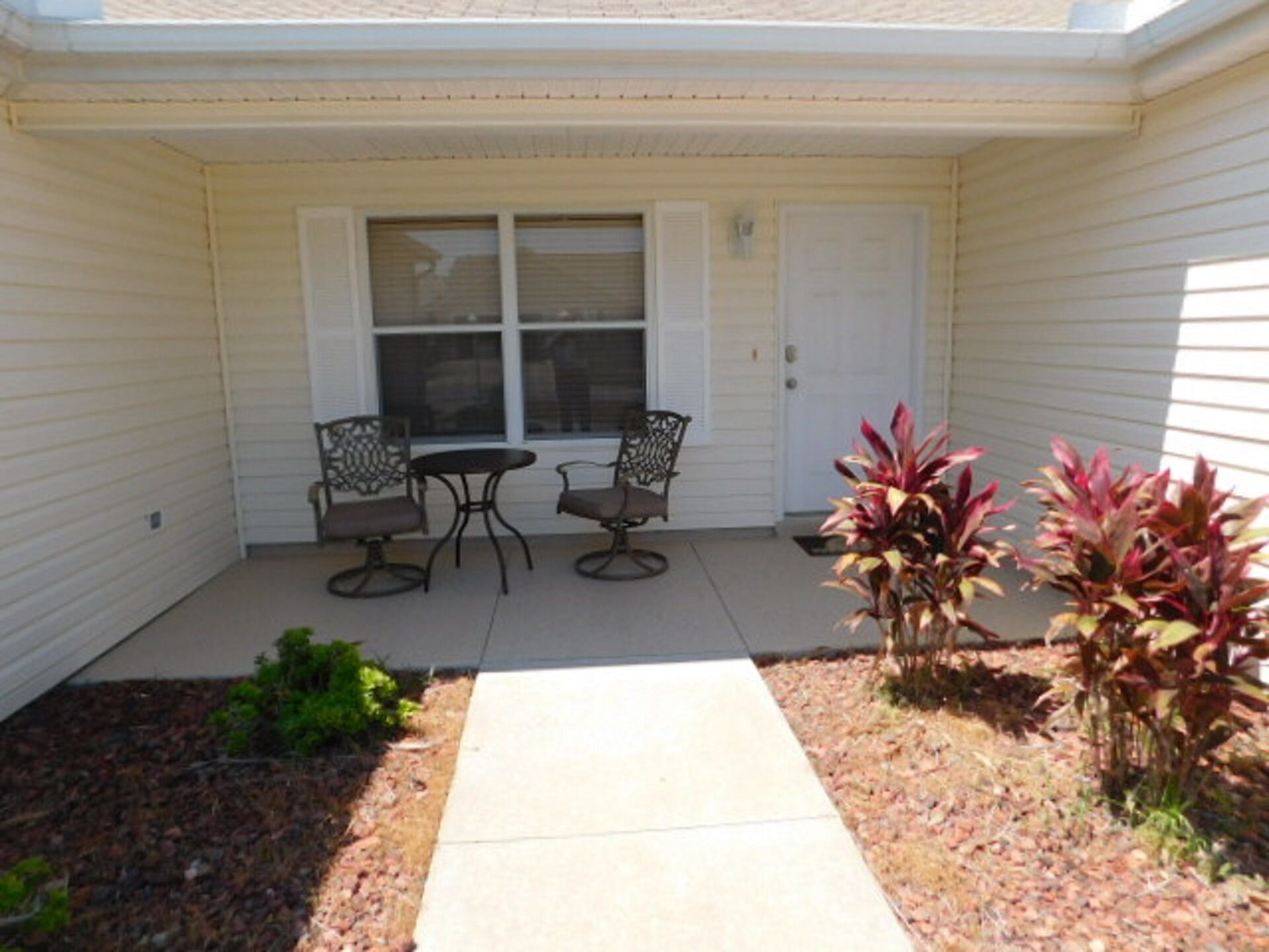 A patio with a table and two chairs, a small garden with plants, and a door with windows.