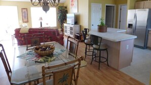 A dining area with a glass table, chairs, and a chandelier. There is a kitchen area with a refrigerator, cabinets, and a countertop.