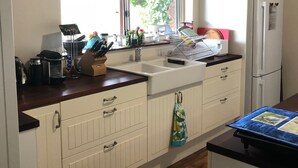 Una cocina con armarios blancos, un lavamanos y una ventana. Hay varios utensilios de cocina en la encimera, incluyendo una cafetera, un hervidor y una licuadora. Se ve un refrigerador al fondo.