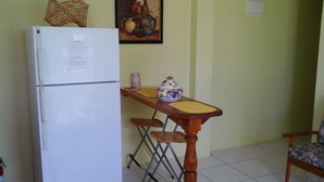 Een compacte keuken met een witte koelkast, een houten tafel met een kom met bloemenmotief, en een opklapbare stoel.