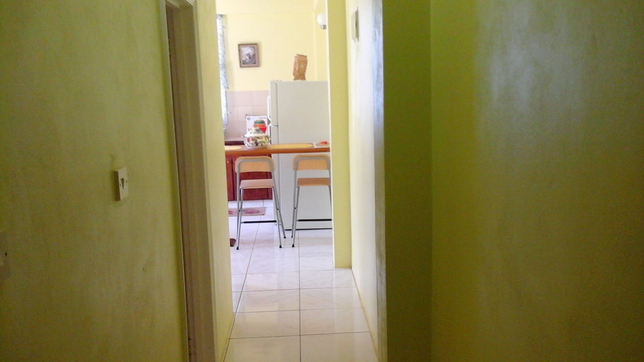 A narrow hallway leading to a kitchen area with a refrigerator, two chairs, and a tiled floor.
