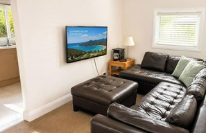 A living room with a flat-screen TV displaying a scenic view, a leather sofa, a wooden side table with a lamp, and a window with blinds.