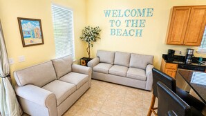 Ein Wohnzimmer mit zwei beigefarbenen Sofas, einem Couchtisch, einem Esstisch mit Stühlen, einer Küchenzeile mit Schränken und Geräten sowie einem an der Wand befestigten Schild mit der Aufschrift 'WELCOME TO THE BEACH'.