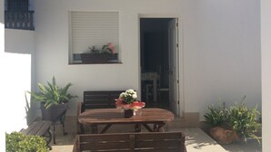 Un patio con una mesa y bancos de madera, plantas en macetas, un jardín colgante con flores y una puerta que da a un espacio interior.