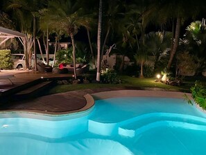 Piscina com deck de madeira, cercada por palmeiras e um carro estacionado por perto.