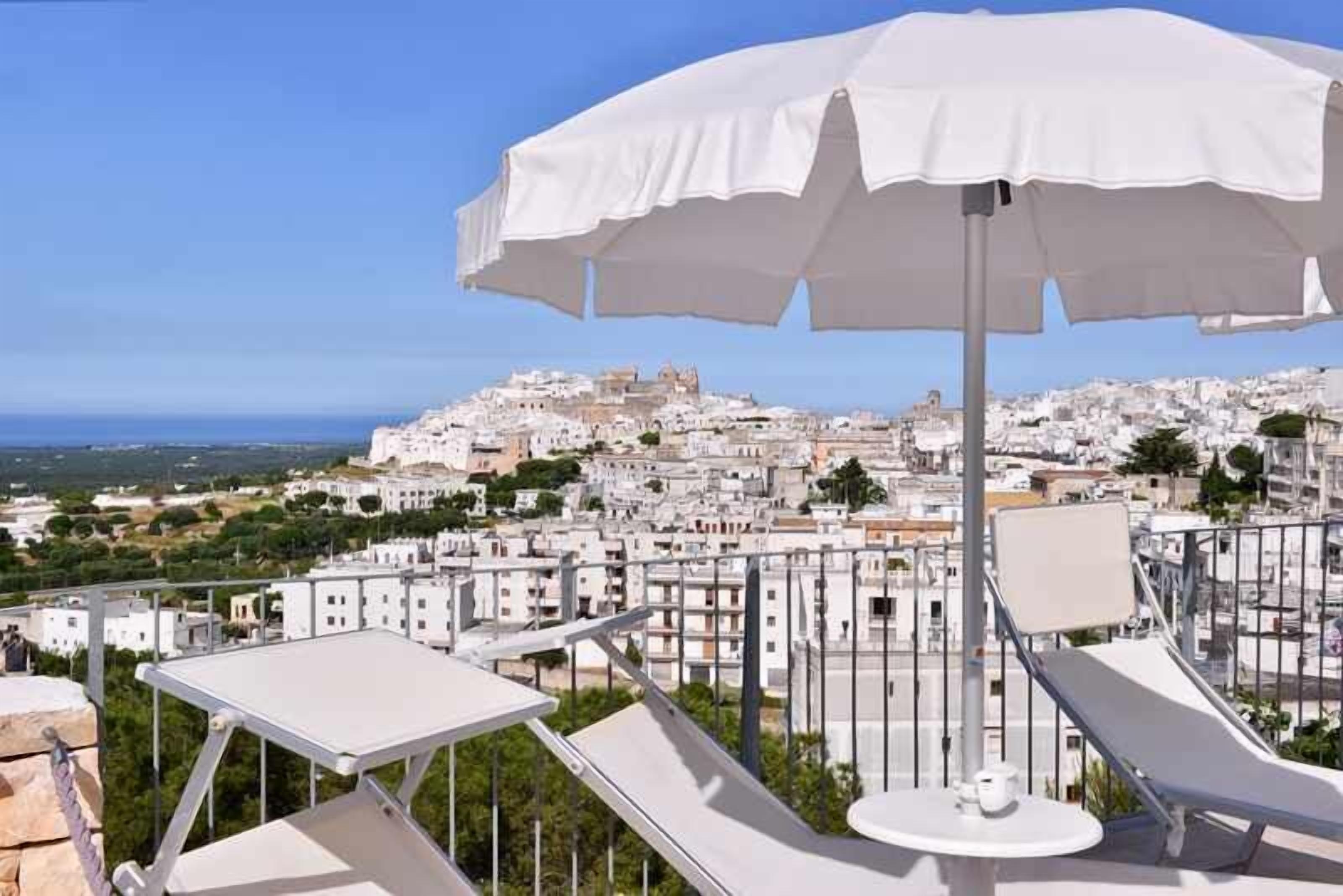 Una terraza en la azotea con sombrillas blancas y sillones de descanso con vistas a un pueblo encalado y al mar.