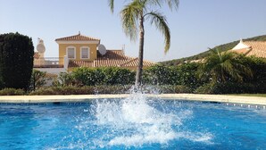 A swimming pool with water splashing, surrounded by a fence, a palm tree, and a building with a tiled roof. - Cozy apartment with beautiful view on golf course, mountains and nature reserve. (Alhaurin El Grande)