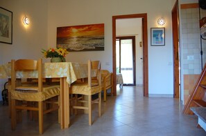 Uma área de refeições com uma mesa posta para quatro pessoas, rodeada por cadeiras de madeira. Há um quadro com um pôr do sol sobre o oceano na parede.