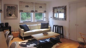 Una sala de estar moderna con un sofá blanco, una mesa de centro y un tapete de cuero de vaca. Hay dos ventanas con cortinas, un espejo y una obra de arte con cebra en la pared.