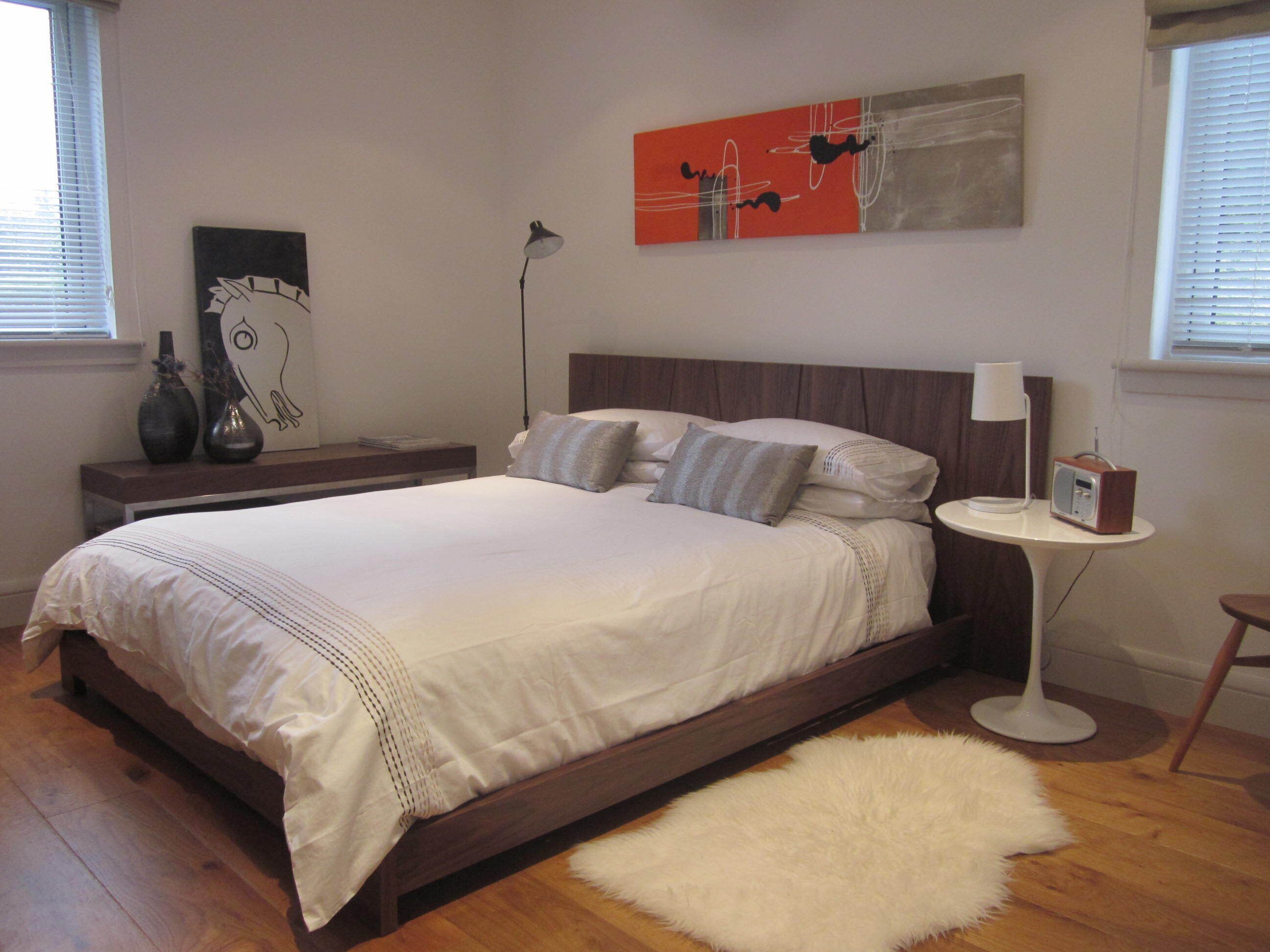 Un dormitorio con cama de madera, ropa de cama blanca, alfombra blanca, mesita de noche con lámpara y radio.