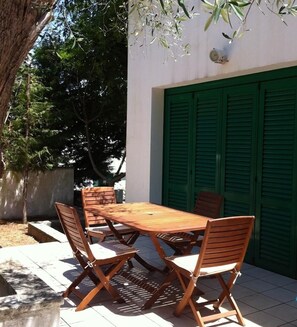 Un patio al aire libre con una mesa de madera y cuatro sillas, una puerta verde y un árbol al fondo.