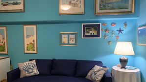 A room with a dark blue sofa, a small round table with a lamp, and various framed pictures on the wall.
