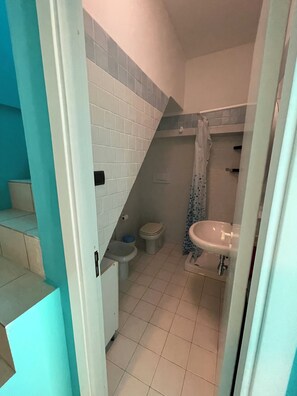 Un baño con paredes de azulejos blancos, un lavamanos blanco, un inodoro y un bidé. Hay una cortina de ducha azul y un cuenco azul. El suelo está cubierto de azulejos.