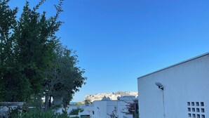 A narrow street with white buildings, greenery, and a distant view of a hill with buildings.